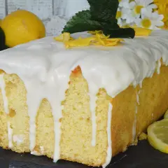 Plumcake al limone glassato, decorato con scorzette gialle e foglioline verdi. Sullo sfondo, limoni freschi e fiori bianchi e gialli.
