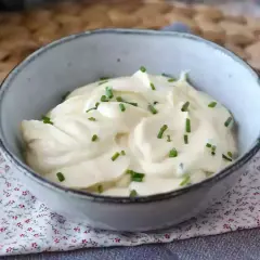 Una cremosa maionese senza uova, guarnita con erba cipollina, in una ciotola grigia su un tovagliolo fiorato.