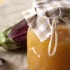 Un vasetto di marmellata di melanzane fatto in casa, con un'melanzana viola sfocata sullo sfondo e un cucchiaino d'argento.