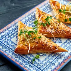 Triangoli dorati di pasta fillo ricetta, cosparsi di semi di sesamo e guarniti con germogli freschi, su un piatto decorato.