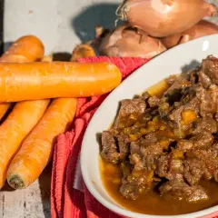 Un invitante spezzatino di manzo in un piatto bianco, con carote fresche e cipolle sullo sfondo.
