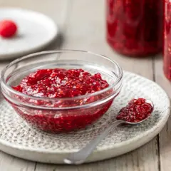 Ciotolina di marmellata di lamponi fatta in casa, con un cucchiaino pieno e lamponi freschi sullo sfondo.