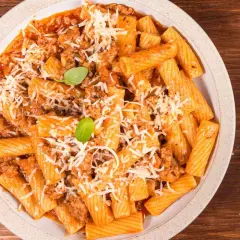 Un piatto di pasta alla zozzona fumante, con rigatoni conditi da un ricco ragù e abbondante parmigiano grattugiato, guarnito con basilico fresco.