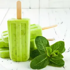 Ghiaccioli alla menta, freschi e verdi, con semi di kiwi visibili, accanto a foglie di menta fresca su un tavolo di legno bianco.