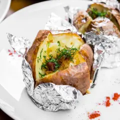 Patate al cartoccio fumanti, condite con erbe fresche e cipolla caramellata, servite su un piatto bianco con un tocco di paprika.