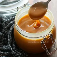 Un cucchiaio raccoglie un filo di caramello salato perfetto, denso e dorato, da un barattolo di vetro.