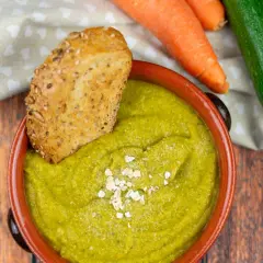 Una ciotola di cremosa crema di zucchine, guarnita con fiocchi d'avena e accompagnata da pane integrale tostato. Carote e zucchine fresche sullo sfondo.