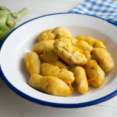 Carciofi fritti alla romana, dorati e croccanti, preparati con una pastella leggera. Accanto, carciofi freschi e un tovagliolo a quadretti.