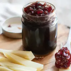 Un vasetto di marmellata di cipolle rosse, con un cucchiaino che ne assaggia un po' su una fetta di formaggio.