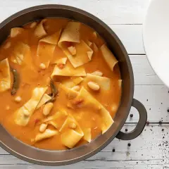 Un saporito piatto di maltagliati con fagioli, servito in una padella scura e una ciotola bianca, con erbe aromatiche e pepe nero a guarnire.