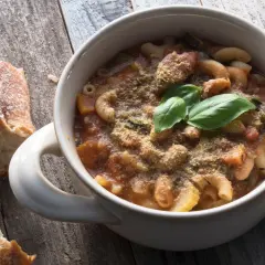 Una ciotola di pasta e fagioli in scatola, guarnita con basilico fresco e parmigiano, accanto a pane rustico su un tavolo di legno.