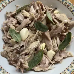 Piatto di tonno di coniglio sfilacciato, condito con aglio e foglie di salvia fresca, servito in una ciotola decorata.