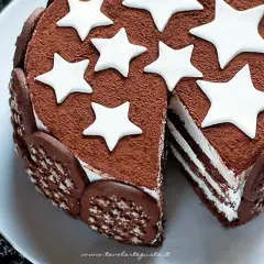 Torta Pan di Stelle ricoperta di cacao, decorata con stelle bianche e biscotti al cioccolato. Una fetta mostra gli strati soffici.