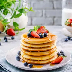 Pila di soffici pancake senza glutine e lattosio, guarniti con fragole, mirtilli e sciroppo d'acero.