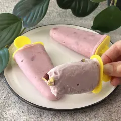 Tre ghiaccioli rosa, un gelato allo yogurt fatto in casa, su un piatto bianco con foglie verdi sullo sfondo.