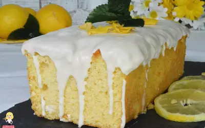 Plumcake al limone glassato, decorato con scorzette gialle e foglioline verdi. Sullo sfondo, limoni freschi e fiori bianchi e gialli.