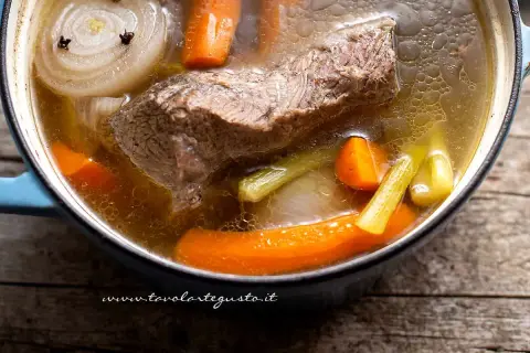 Brodo di carne ricetta: un pezzo di carne, carote, sedano e cipolla con chiodi di garofano bollono in una pentola.