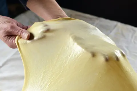 Mani che stendono la pasta matta, seguendo la ricetta originale. La pasta è sottile e traslucida, con le dita che la modellano.