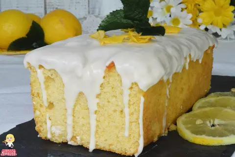 Plumcake al limone glassato, decorato con scorzette gialle e foglioline verdi. Sullo sfondo, limoni freschi e fiori bianchi e gialli.