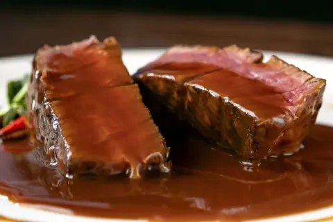 Succulento filetto di manzo in padella, tagliato a fette e nappato con una ricca salsa bruna, servito su un piatto bianco con contorno verde.