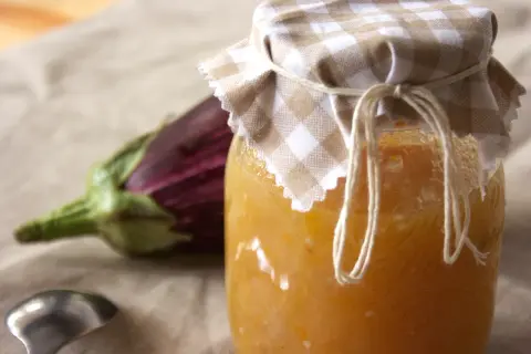 Un vasetto di marmellata di melanzane fatto in casa, con un'melanzana viola sfocata sullo sfondo e un cucchiaino d'argento.