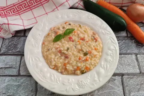 Un cremoso risotto con verdure, guarnito con basilico fresco. Accanto, carote, zucchine e cipolla, ingredienti per un piatto gustoso.