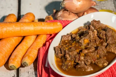 Un invitante spezzatino di manzo in un piatto bianco, con carote fresche e cipolle sullo sfondo.