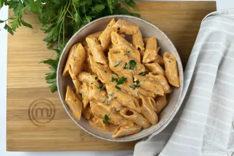 Penne alla vodka cremose e invitanti, guarnite con prezzemolo fresco, su un tagliere di legno con il logo MasterChef.