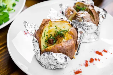 Patate al cartoccio fumanti, condite con erbe fresche e cipolla caramellata, servite su un piatto bianco con un tocco di paprika.