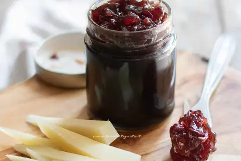 Un vasetto di marmellata di cipolle rosse, con un cucchiaino che ne assaggia un po' su una fetta di formaggio.
