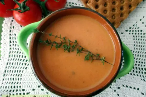 Una ciotola di cremosa crema di pomodoro, guarnita con timo fresco, accanto a pomodori maturi e cracker croccanti.