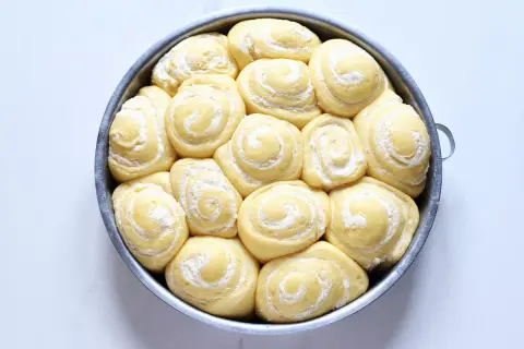 Torta di rose lievitata, pronta per essere infornata, con soffici spirali di pasta e una spolverata di zucchero a velo.