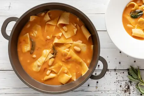 Un saporito piatto di maltagliati con fagioli, servito in una padella scura e una ciotola bianca, con erbe aromatiche e pepe nero a guarnire.