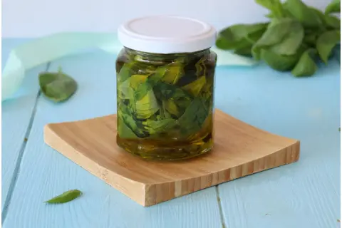 Un barattolo di olio al basilico fatto in casa, con foglie fresche immerse nell'olio dorato, su un tagliere di legno.