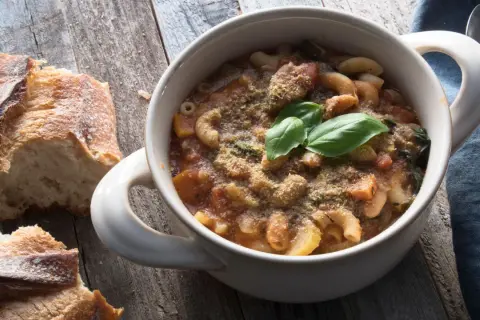 Una ciotola di pasta e fagioli in scatola, guarnita con basilico fresco e parmigiano, accanto a pane rustico su un tavolo di legno.