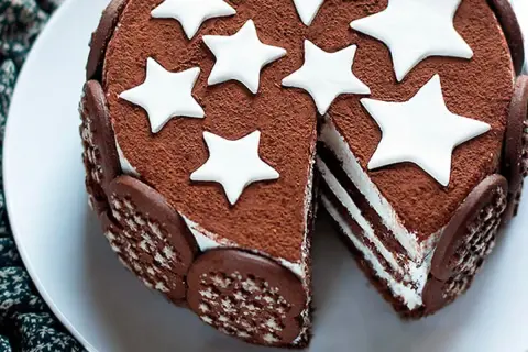 Torta Pan di Stelle ricoperta di cacao, decorata con stelle bianche e biscotti al cioccolato. Una fetta mostra gli strati soffici.