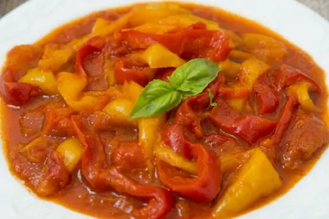 Peperonata ricetta della nonna: peperoni rossi e gialli cotti lentamente in un sugo saporito, guarniti con una fogliolina di basilico fresco.