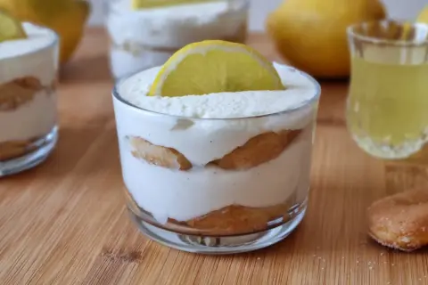 Delizioso tiramisù al limoncello in bicchieri, guarnito con una fetta di limone. Accanto, un bicchierino di limoncello e savoiardi.