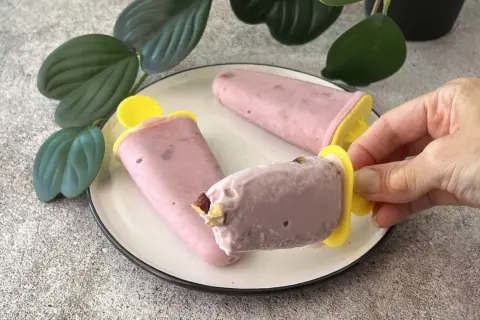 Tre ghiaccioli rosa, un gelato allo yogurt fatto in casa, su un piatto bianco con foglie verdi sullo sfondo.