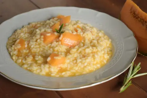 Un cremoso risotto con pezzi di melone e un rametto di rosmarino, accanto a una fetta di melone.
