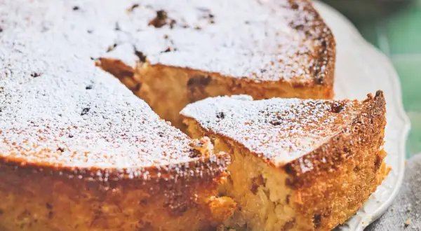 Torta di riso ricetta originale, soffice e dorata, cosparsa di zucchero a velo. Una fetta mostra la consistenza cremosa.