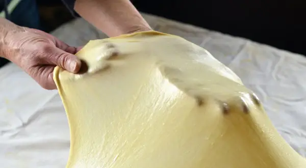 Mani che stendono la pasta matta, seguendo la ricetta originale. La pasta è sottile e traslucida, con le dita che la modellano.