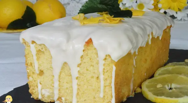 Plumcake al limone glassato, decorato con scorzette gialle e foglioline verdi. Sullo sfondo, limoni freschi e fiori bianchi e gialli.