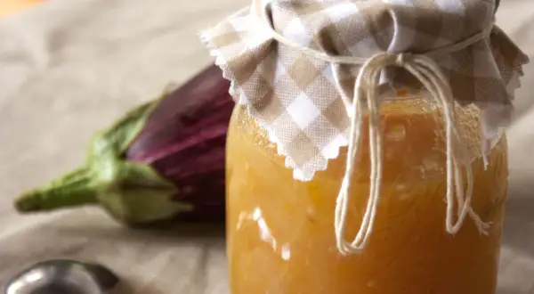 Un vasetto di marmellata di melanzane fatto in casa, con un'melanzana viola sfocata sullo sfondo e un cucchiaino d'argento.