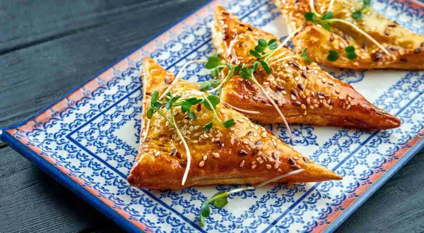 Triangoli dorati di pasta fillo ricetta, cosparsi di semi di sesamo e guarniti con germogli freschi, su un piatto decorato.