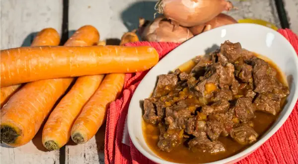Un invitante spezzatino di manzo in un piatto bianco, con carote fresche e cipolle sullo sfondo.