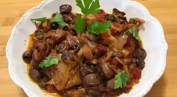 Un piatto invitante di melanzane in agrodolce, con olive nere e capperi, guarnito con prezzemolo fresco.