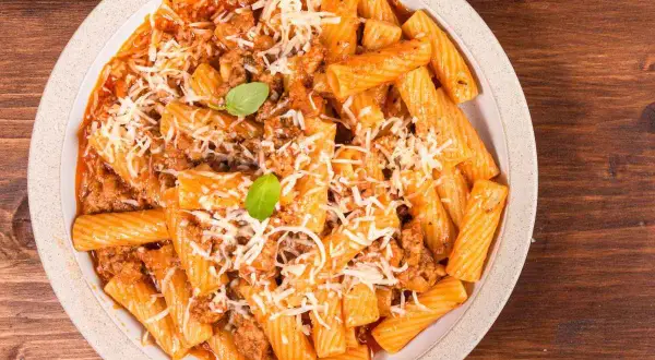 Un piatto di pasta alla zozzona fumante, con rigatoni conditi da un ricco ragù e abbondante parmigiano grattugiato, guarnito con basilico fresco.