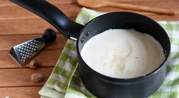 Besciamella senza glutine cremosa in pentolino, con noce moscata grattugiata. Accanto, noce moscata intera e una grattugia.
