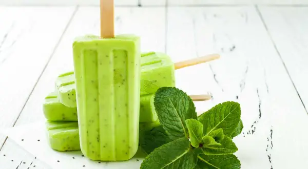 Ghiaccioli alla menta, freschi e verdi, con semi di kiwi visibili, accanto a foglie di menta fresca su un tavolo di legno bianco.